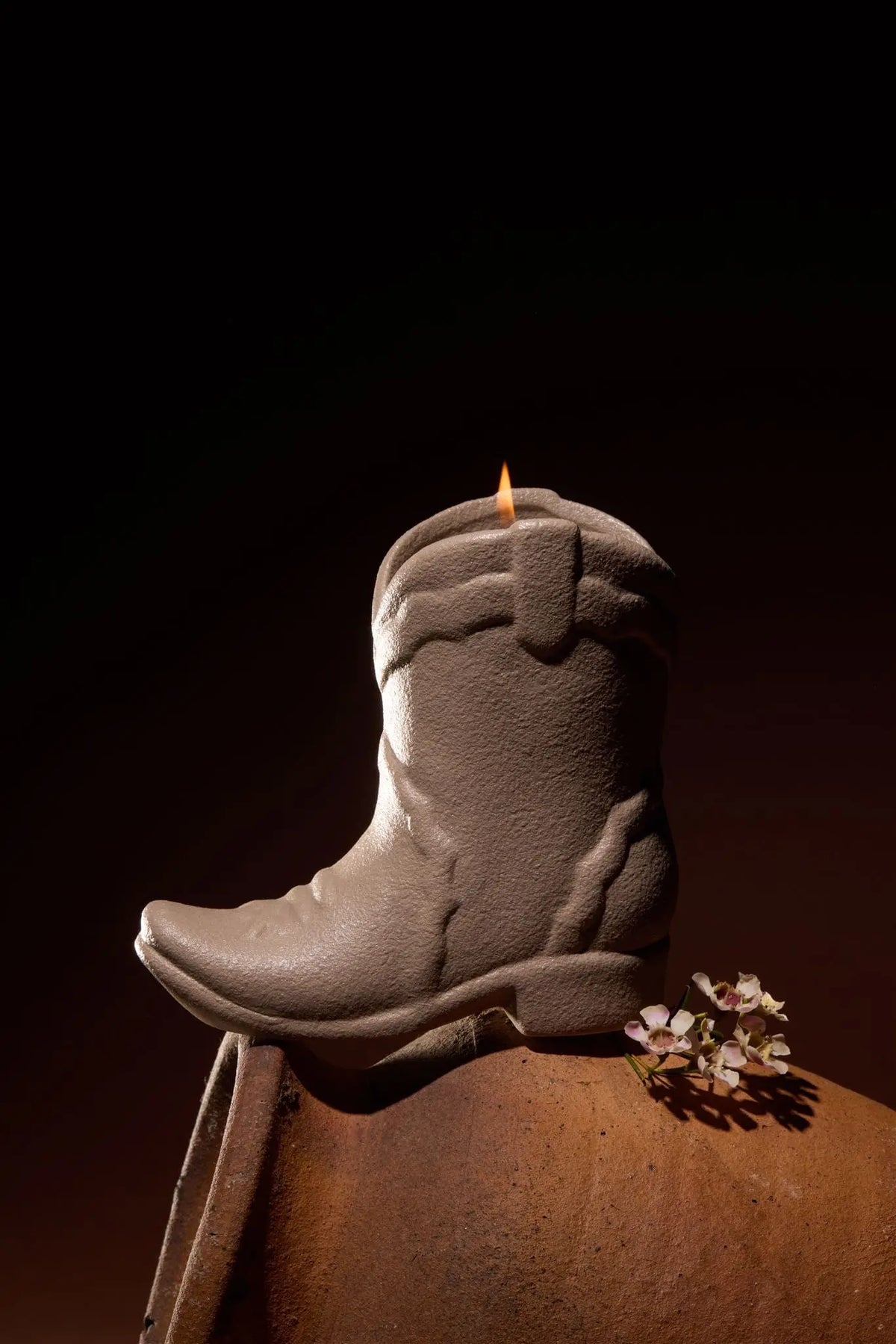 A Bergamot & Mahogany - Nashville Boot 6 oz Candle, shaped like a gray cowboy boot with a gentle flame atop, rests on a brown surface. Beside it, small dried flowers evoke the rustic charm of Nashville-inspired décor and the inviting warmth of western fragrances against a dark background.
