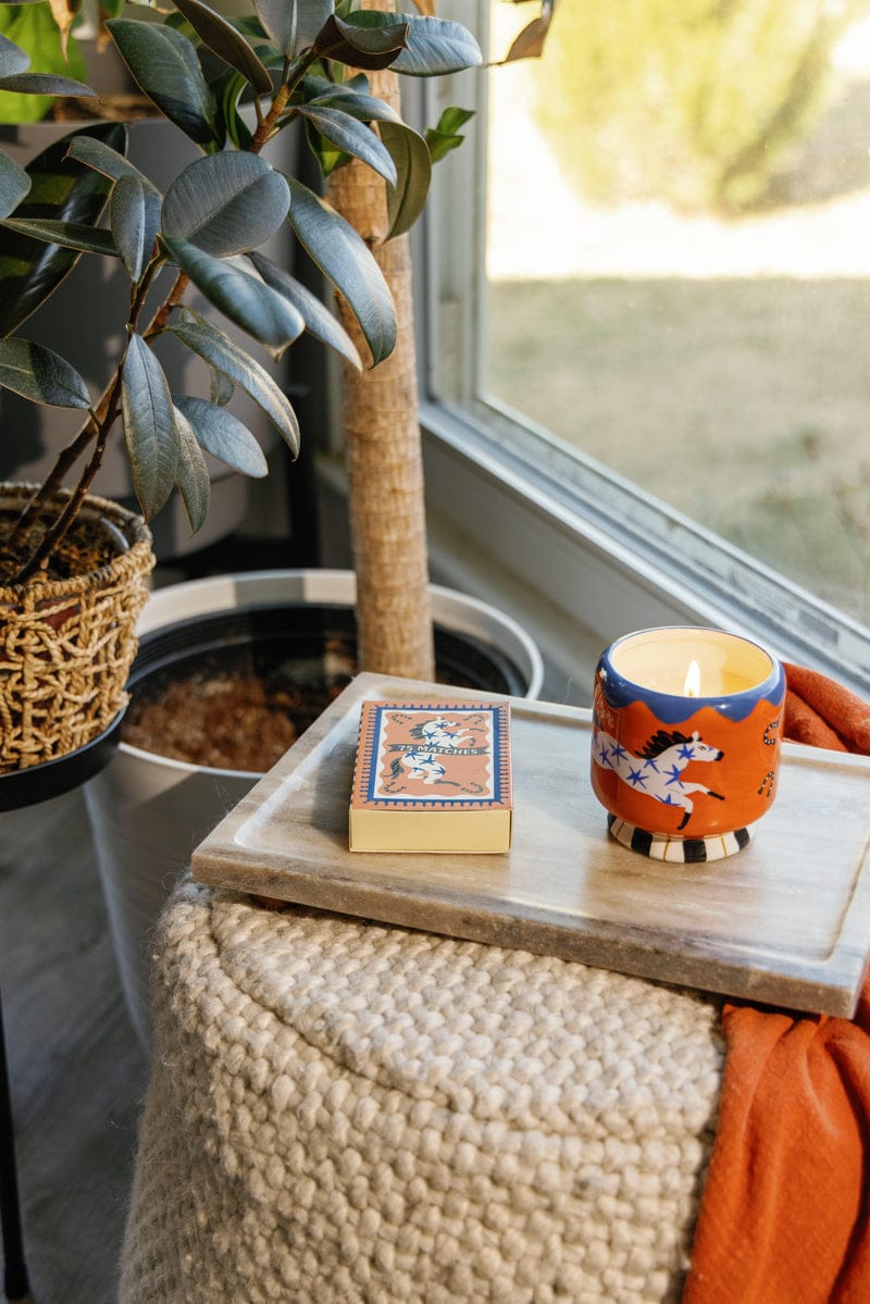 The Sienna Sunset - A Dopo 8 oz Candle rests on a marble tray beside a potted plant and knit blanket by the window, paired with a matching box of matches for a cozy touch.