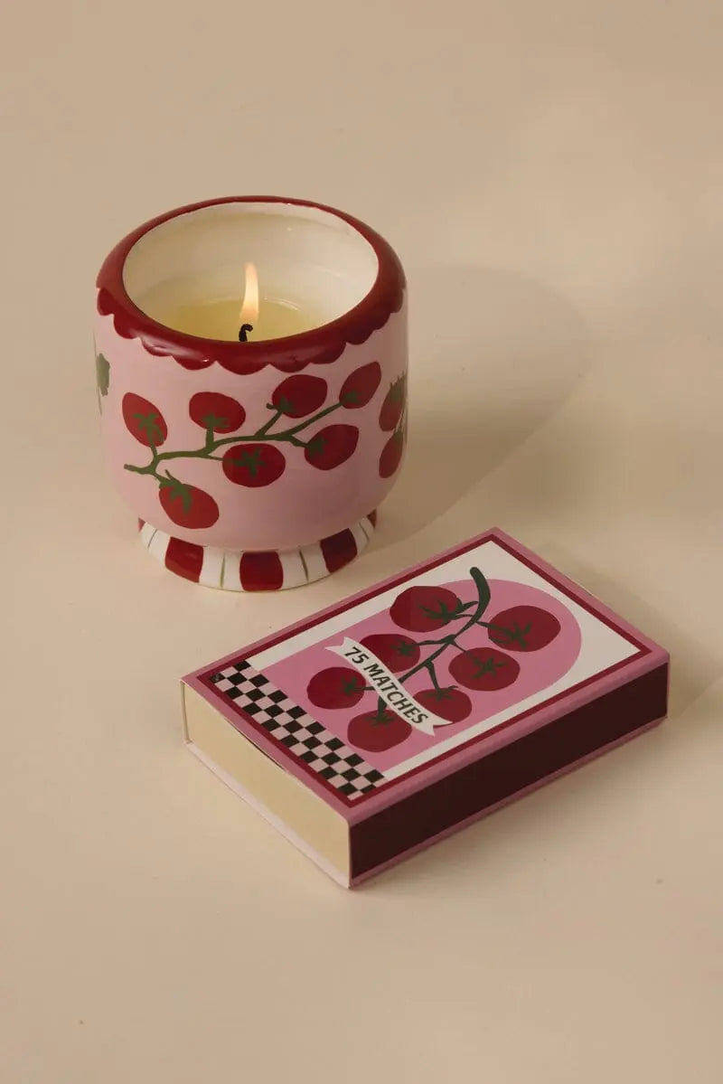 An A Dopo candle in a pink holder with red cherry designs sits next to an A Dopo "Tomato Vine" 75ct boxed matches, both sharing the same cheerful pattern reminiscent of Mediterranean travel, on a beige surface.