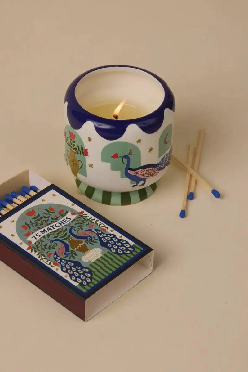 An A Dopo "Peacock" candle in a decorative holder sits next to an open box of A Dopo "Peacock" 75ct boxed matches and two loose matches on a beige surface.