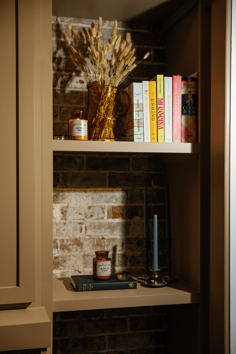 Two shelves with books, the Saffron + Rose - Apothecary 8 oz Candle in a glass vessel, dried wheat in a glass vase, and a brass candleholder with a blue candle, set against an exposed brick wall background.