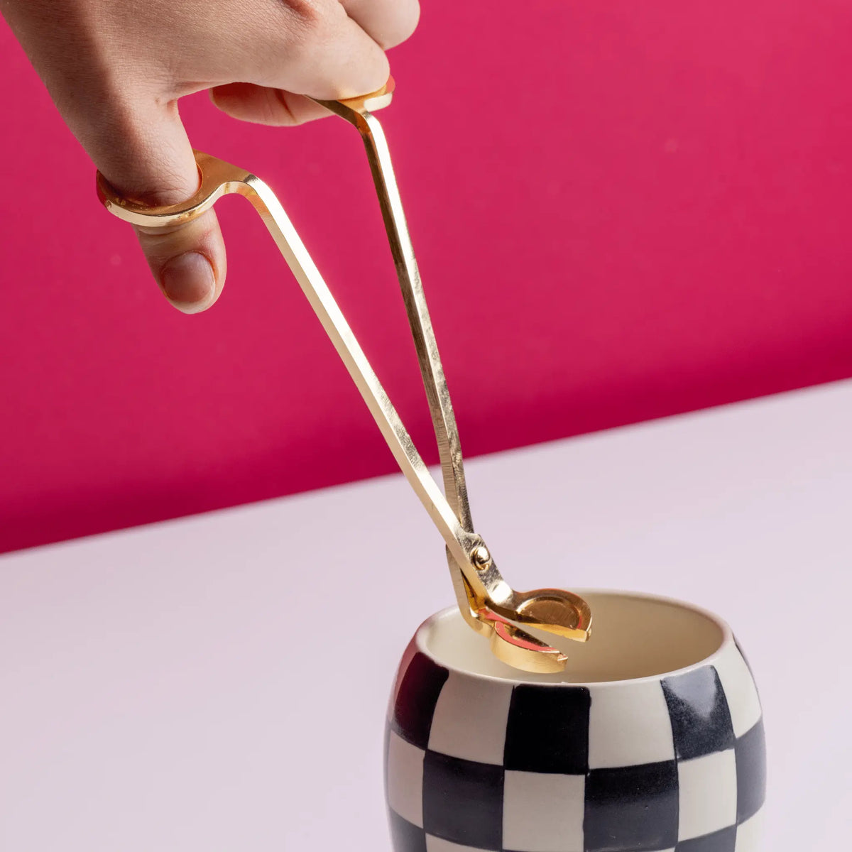 A hand holds the Wick Trimmer above a black-and-white checkered cup, set against a pink background on a white surface—a stylish candle accessory scene.
