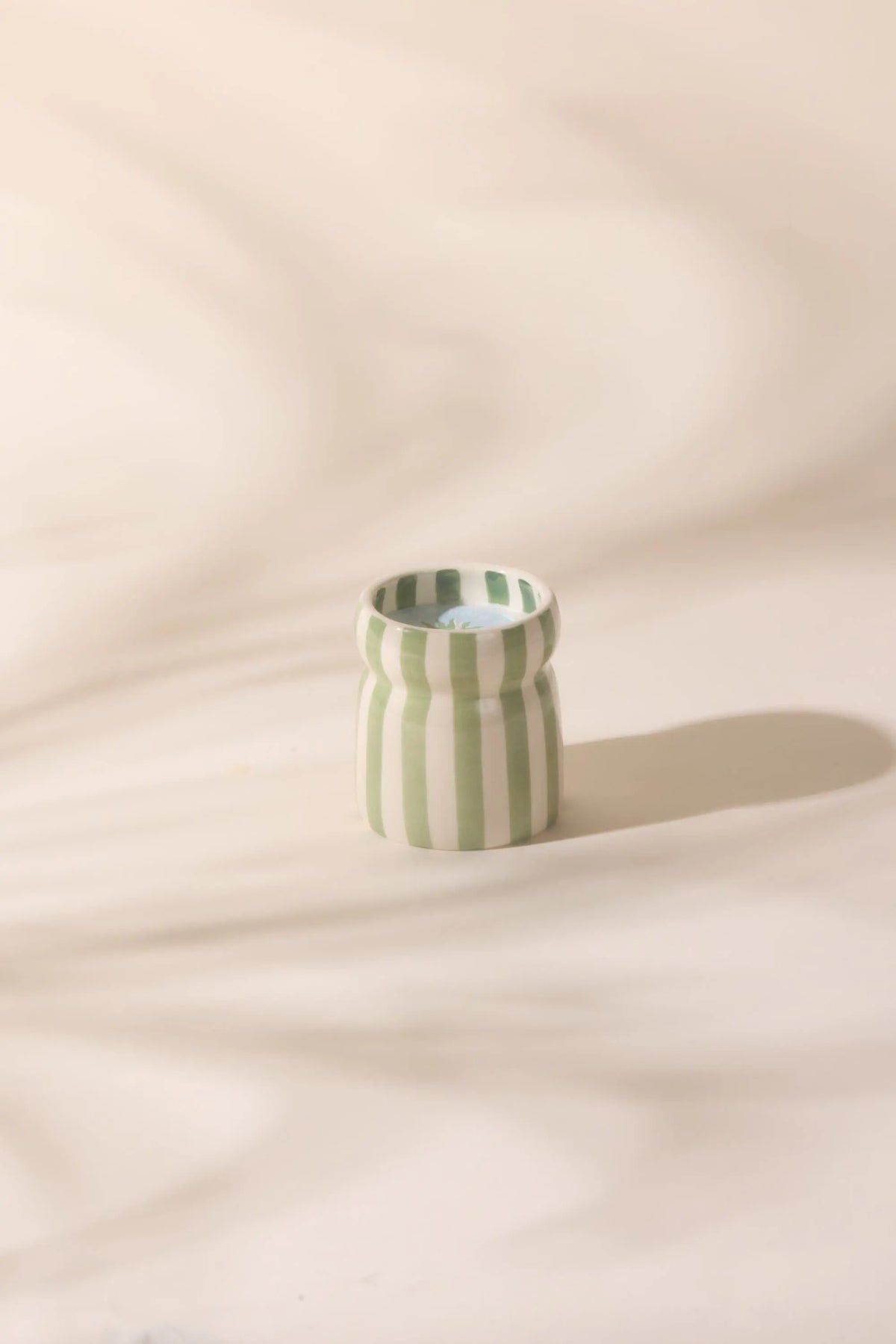 The Bora Bora - Cabana 6.5 oz Candle, in a small ceramic cup with vertical green and white stripes, sits on a light surface accented by soft background shadows.