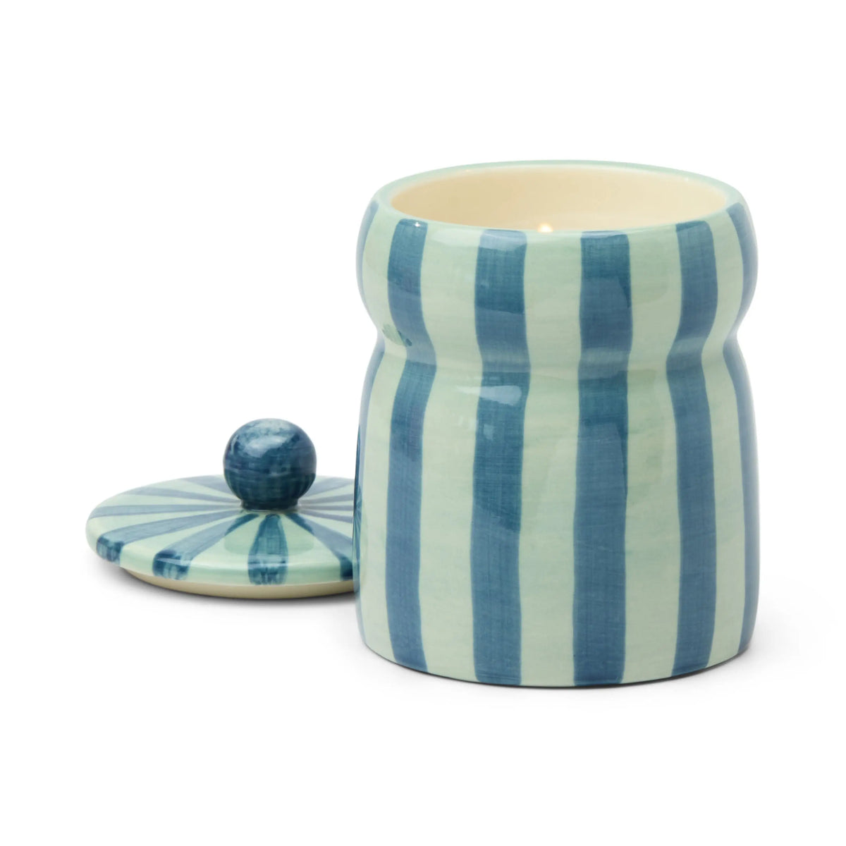 Presented on a plain white background, the Warm Cider - Cabana 6.5 oz Candle is housed in a hand-painted ceramic vessel adorned with vertical blue and white stripes. The matching lid, topped with a round knob, is placed beside the jar, showcasing the elegant design of this unique candle container.