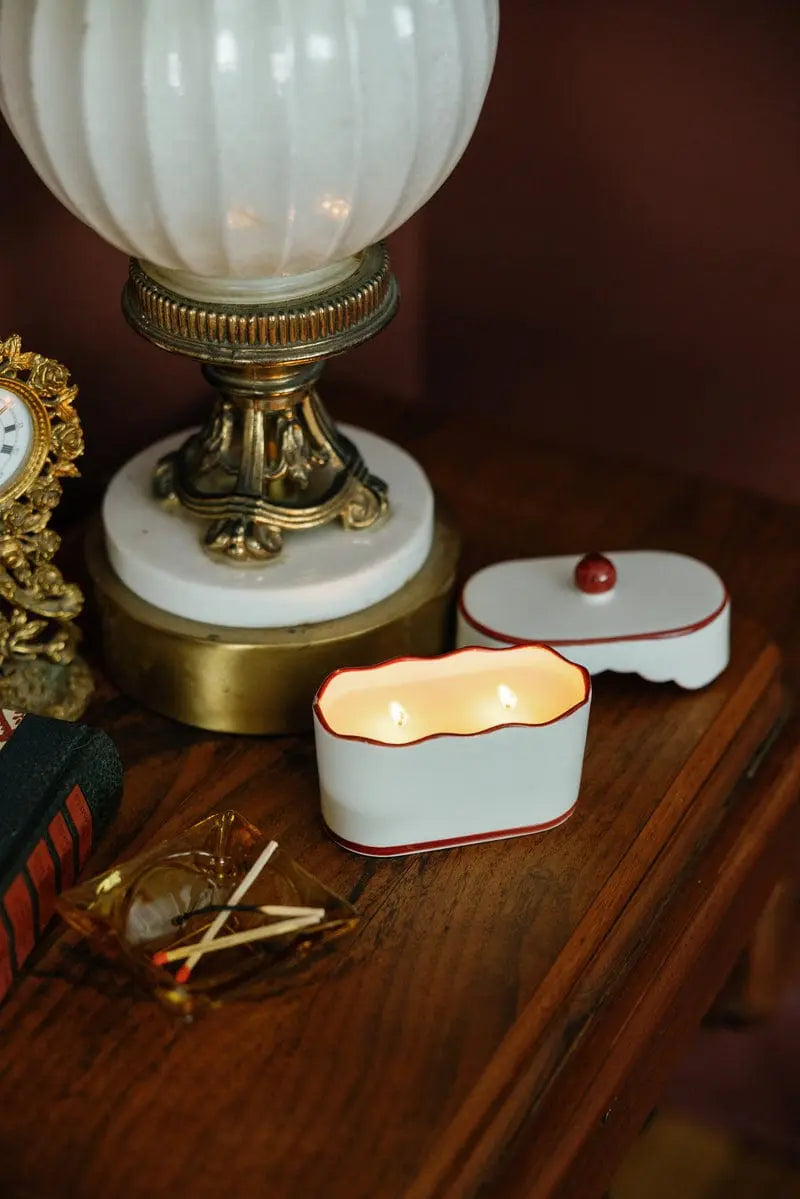 A lit French Linen & Orris - Chérie 5.5 oz Ceramic Candle with two wicks sits in a porcelain vessel on a wooden table near a lamp, an open lid, a small clock, and a glass tray holding several matches.
