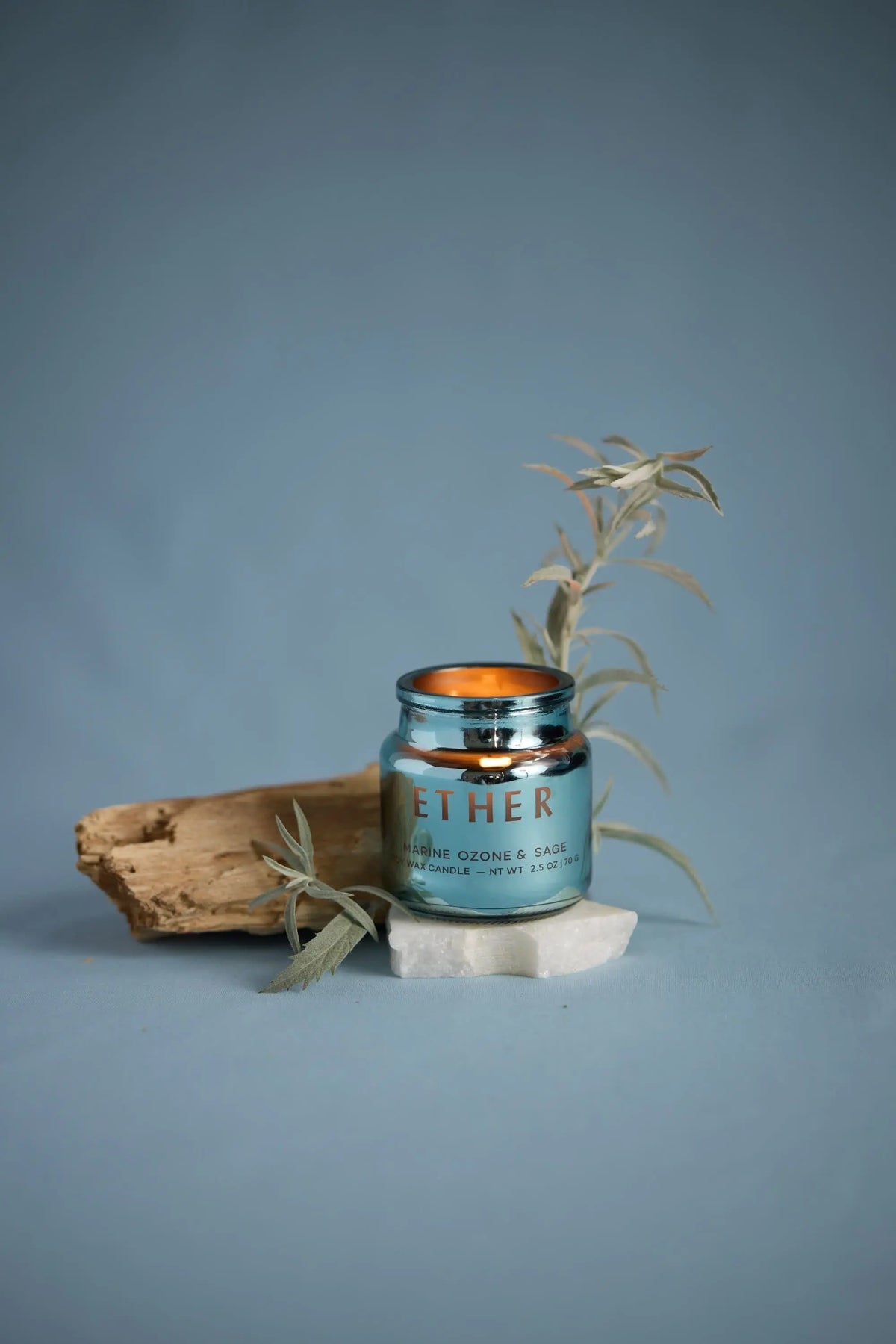 A blue Sea Salt & Sage - Chroma 2.5 oz candle jar sits on a white stone with a beige wood piece and a light green leafy twig beside it, set against a plain blue background. Part of the Chroma collection, this candle is housed in electroplated glass for an elegant display.