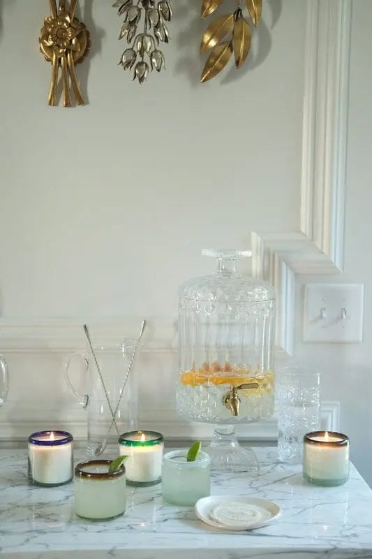 A marble countertop displays the Vanilla Rosa - La Playa 9 oz Candle, a glass dispenser with citrus-infused water, empty glasses, and decorative gold wall accents.