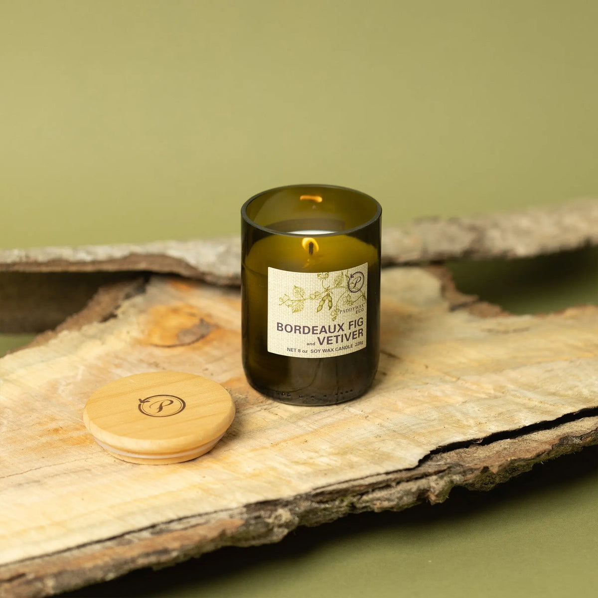 The Bordeaux Fig + Vetiver - Eco 8 oz Candle glows in a green recycled vessel on a wooden board, its logo-engraved wooden lid nearby. Olive green bark pieces enhance the earthy setting.
