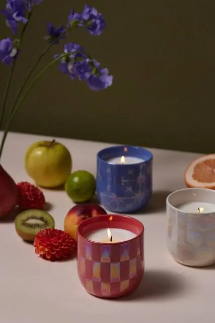 Three lit candles, including the Sapphire Waters - Lustre Candle 10 oz in a patterned glass holder, are arranged on a table with fresh fruit, flowers, and a neutral-colored background.