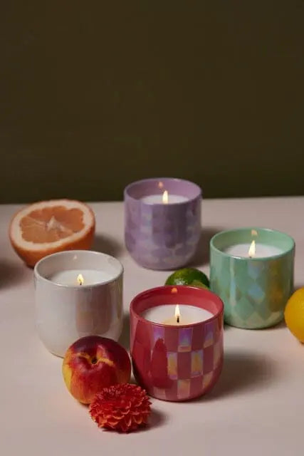 Four lit Lavender & Fern - Lustre Candle 10 oz luxury scented candles in colorful, patterned jars are arranged on a surface with peach, grapefruit, lime, lemon, and a red carnation flower nearby.