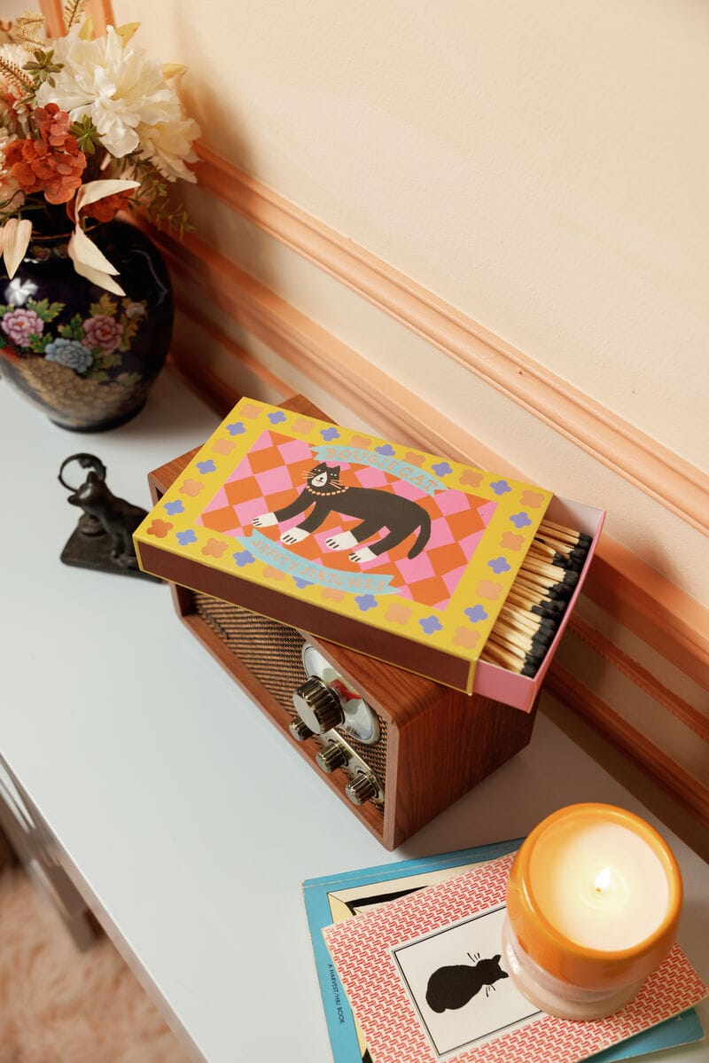 A colorful Bougie Cat - Large Rectangle Boxed Matches with a cat illustration sits on a radio beside a candle, books, figurine, and floral vase—perfect decor for cat lovers and match enthusiasts.