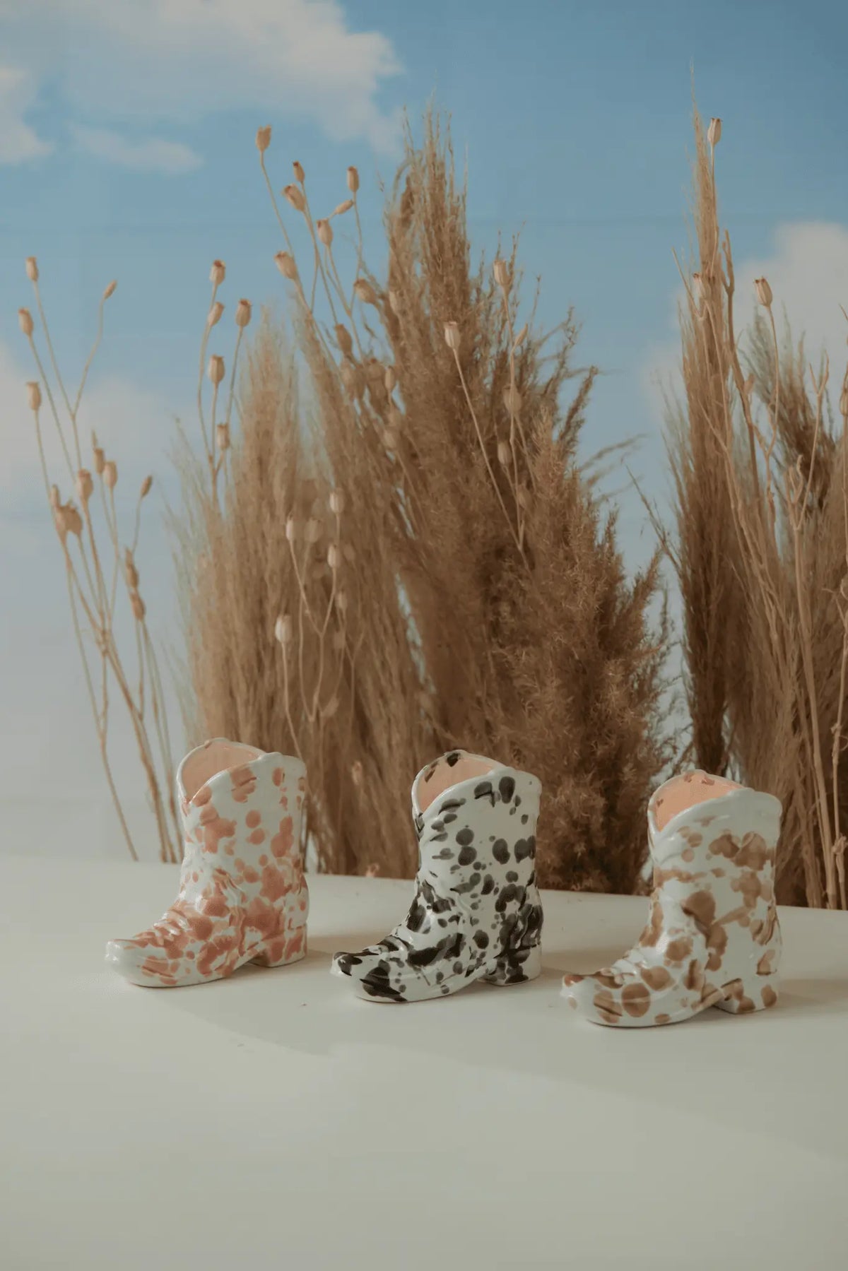 Three small Nashville Boot 6 oz candles in Palo Santo Suede scent, with pink, black, and brown cow-print patterns, are displayed on a white surface with tall dried grasses and a blue sky backdrop for a Western-inspired vibe.