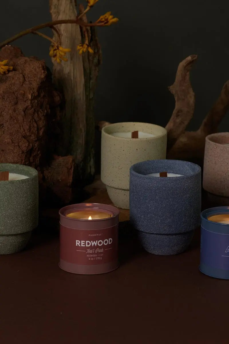 The Glacier - Parks 11 oz Candle with a crackling wood wick sits on a brown surface amid wood and rocks, evoking the scents and textures of national parks.