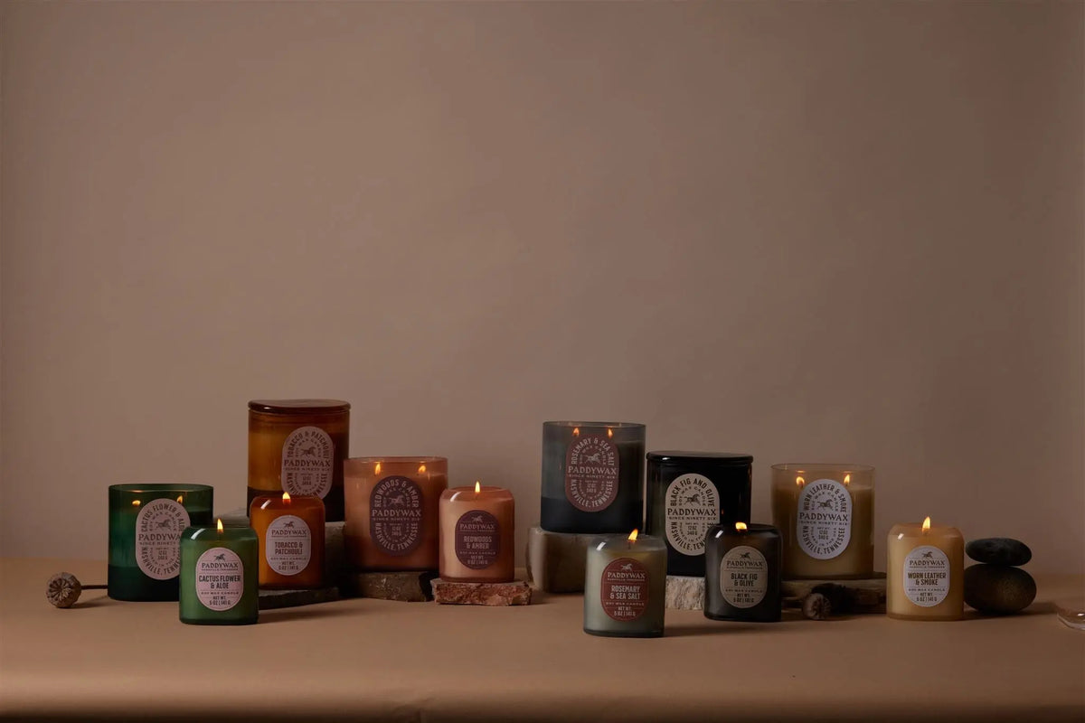 A Redwoods + Amber - Vista 12 oz Candle, designed in a vintage-inspired glass jar, is arranged on a table among stones and wooden pieces. The elegant candle, part of the Vista collection, stands out against a plain beige background.