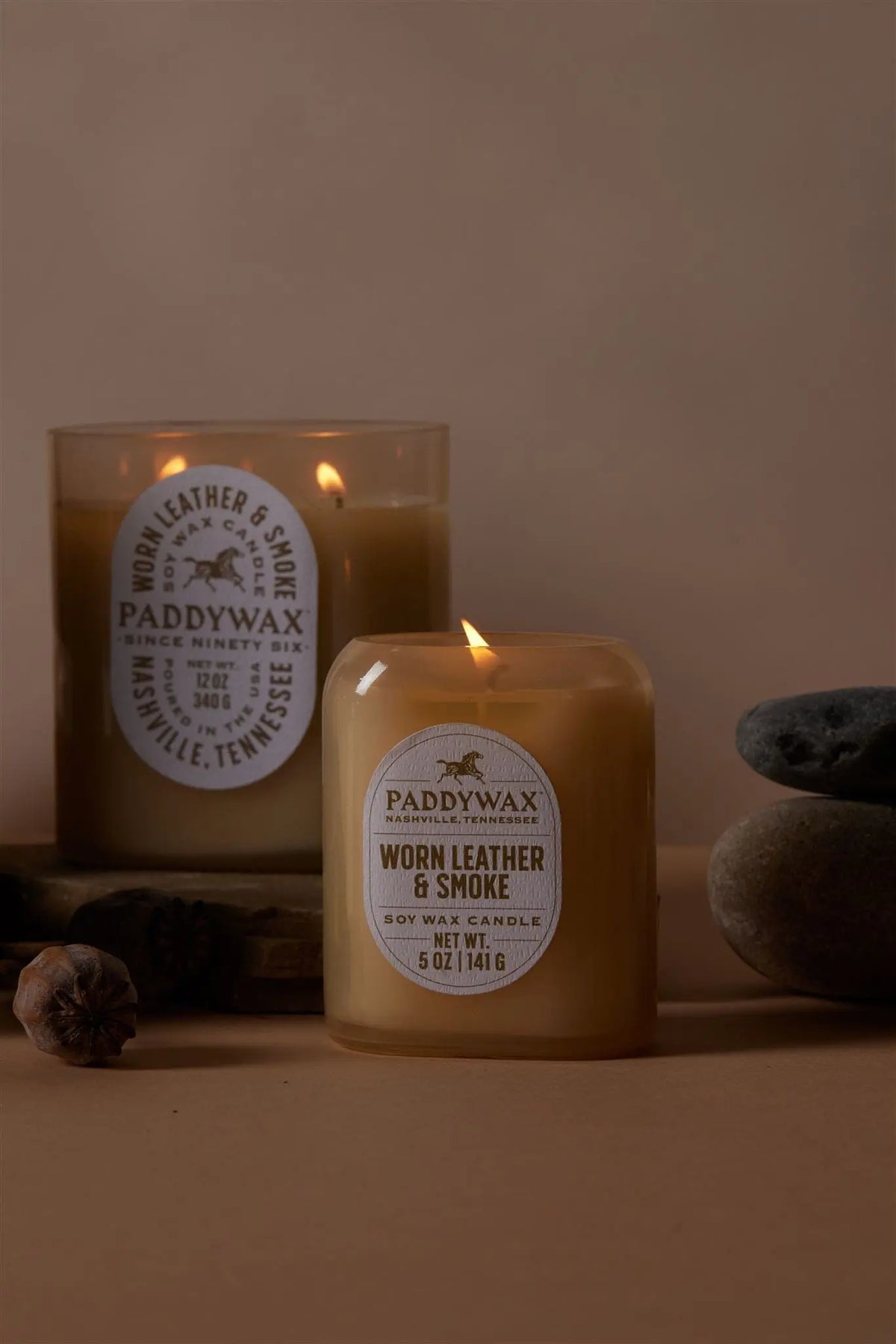 Two Worn Leather & Smoke - Vista 5 oz Candles are shown on a beige surface, one lit and one unlit, with gray stones and a dried seed podโideal for modern decor and adding a luxurious fragrance to your home.