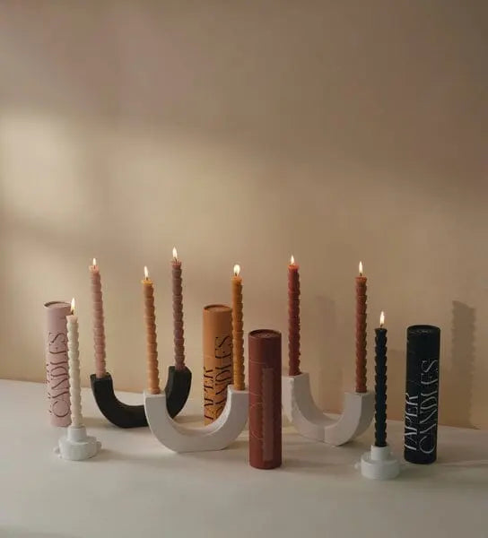 Seven spiral-designed candles are arranged in modern black and white candle holders, featuring the striking U Taper Holder - Black. Among them, three cylindrical containers labeled UVAT CANDLES sit on a neutral surface set against a beige wall, adding an elegant touch of minimalist décor.