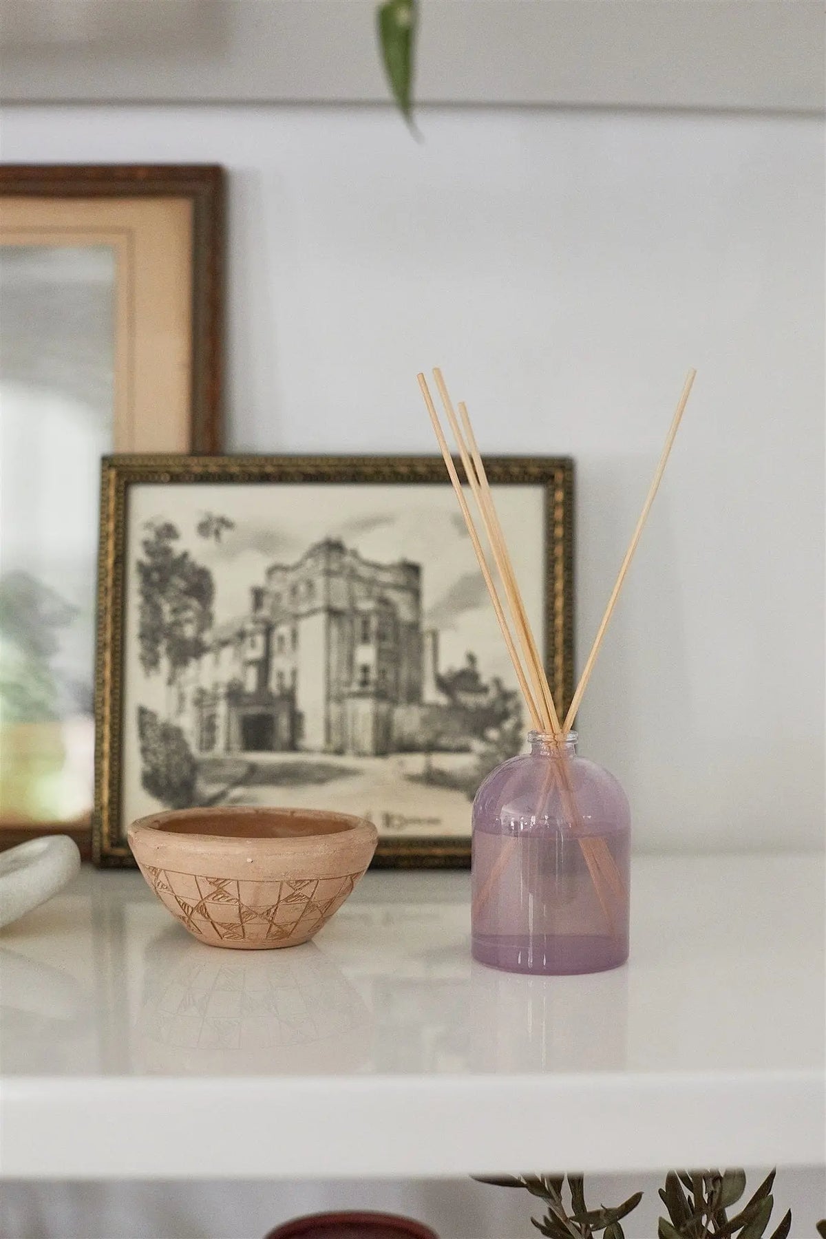 A small beige bowl and a Lavender - 1.5 oz Diffuser with a calming lavender scent sit on a white shelf in front of a framed black-and-white architectural drawing.