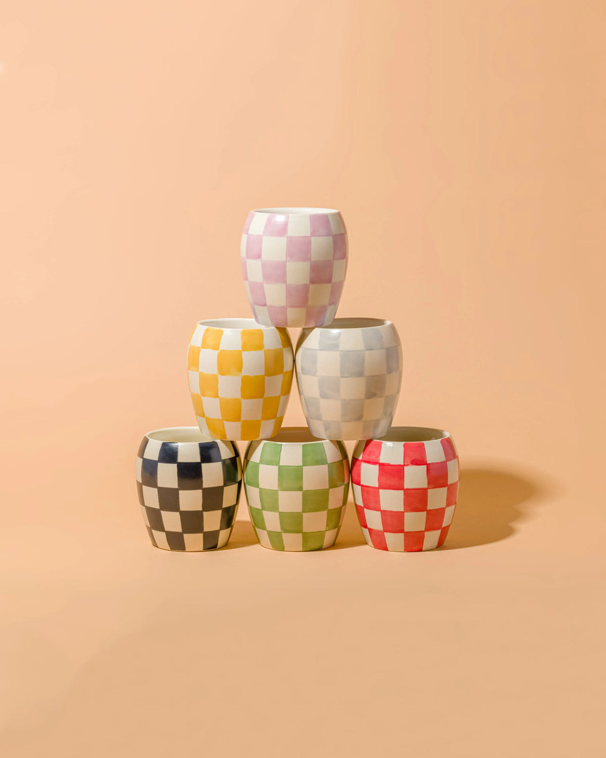 Five Checkmate 11 oz Candles in Cactus Flower are stacked in a pyramid against a peach background. These hand-painted vessels display black, green, red, yellow, pink, and blue checkered patterns, adding a playful touch to your reusable decor.