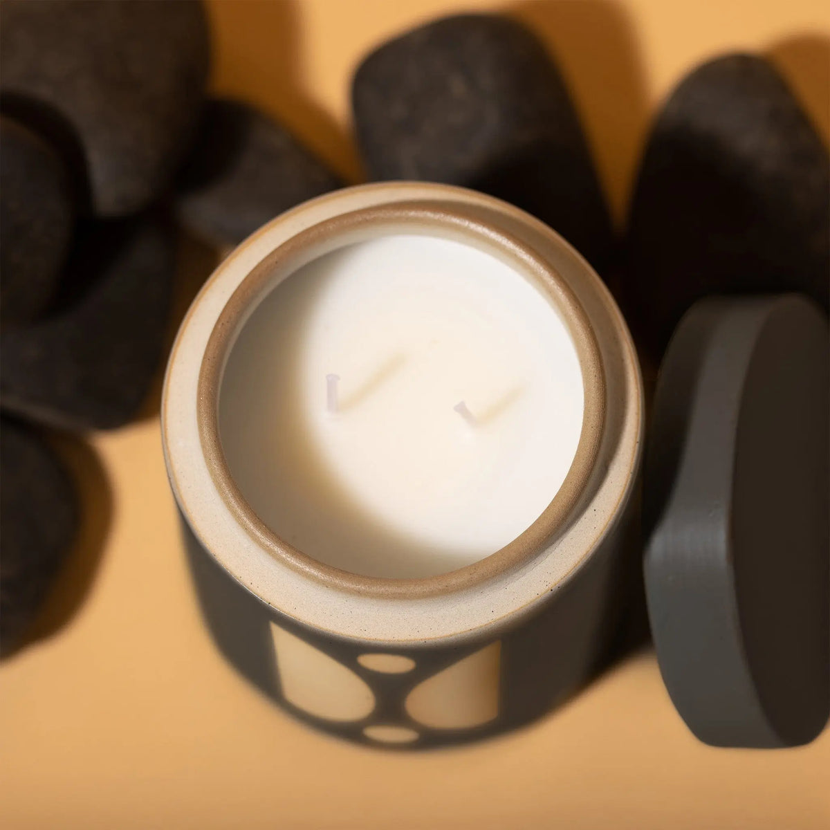 A top view of the unlit Palo Santo Suede - Form 12 oz Candle with two wicks in a decorative container, placed beside smooth black stones on a beige surface.