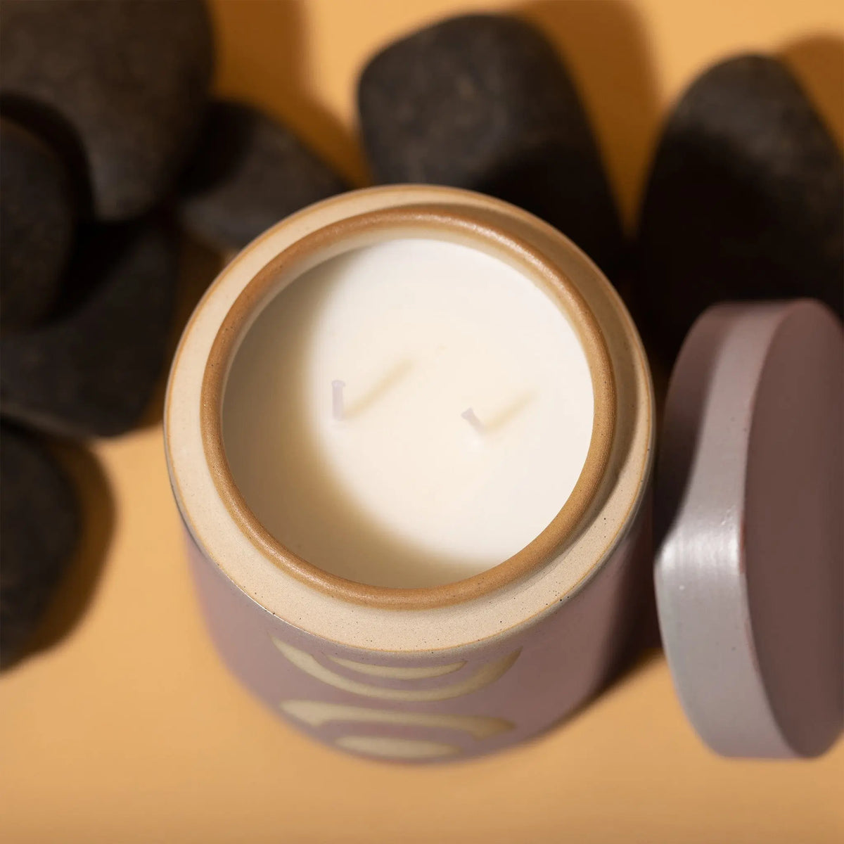 A top view of the Mandarin Mango - Form 12 oz Candle, a white ceramic two-wick candle in a round container with the lid set aside, displayed beside smooth black stones on a beige surface for a tranquil home decor touch.
