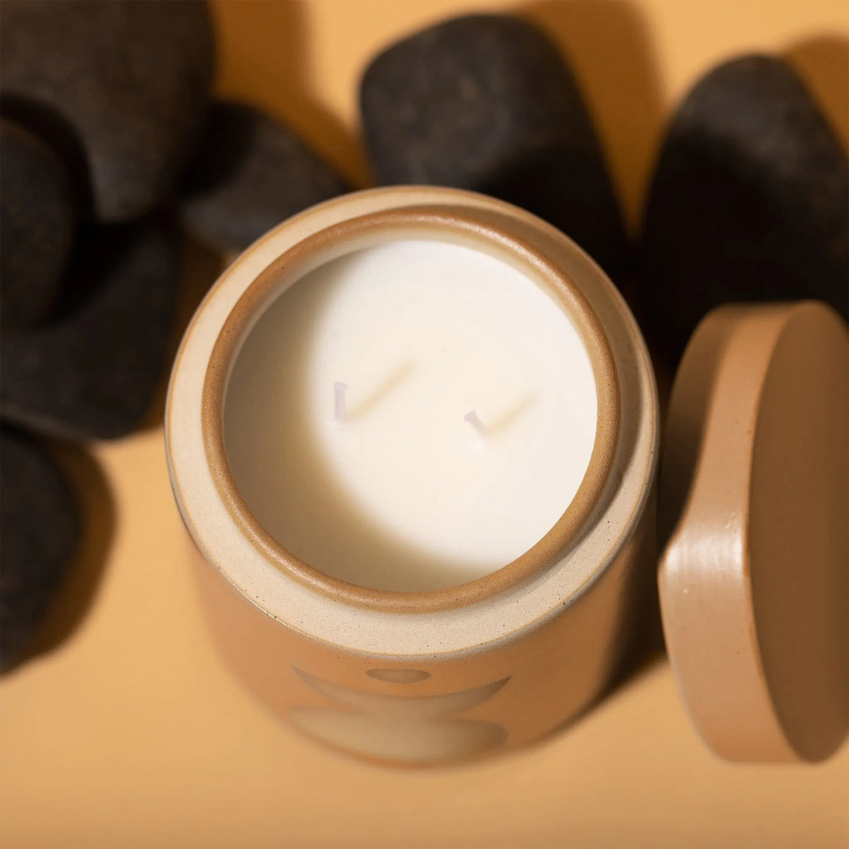 The Wild Fig + Vetiver - Form 12 oz Candle is a beige ceramic candle with two wicks, displayed without the lid and black stones nearby, set on a light brown surface.