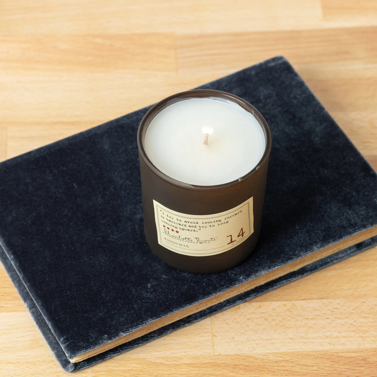 The Charlotte Bronte - Library Candle 6 oz, a white literary candle in a dark glass jar, sits atop a closed dark blue velvet-covered book on a light wooden surface—an ideal fragrance gift from the Library Collection.
