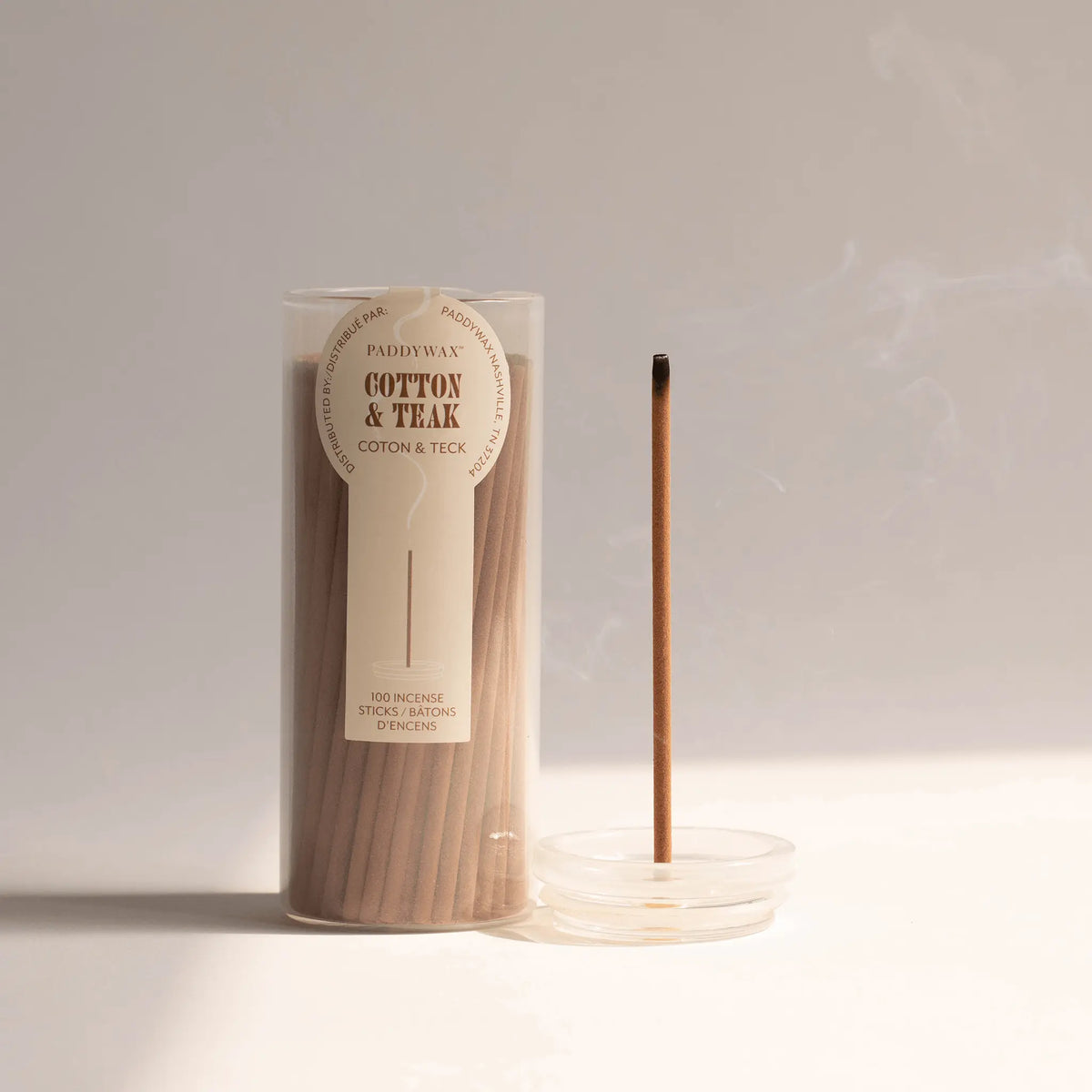 A clear container labeled “Cotton + Teak - Haze Incense” showcases a refined collection of incense sticks. Nearby, a single stick stands upright in an incense holder, releasing a delicate, smokeless aroma against a neutral backdrop.