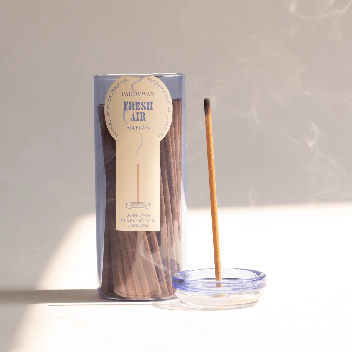 A clear container labeled "Fresh Air - Haze Incense" holds a collection of slender incense sticks. One stick is burning in a small glass incense holder, its flameless fragrant smoke curling in soft sunlight across a light surface.