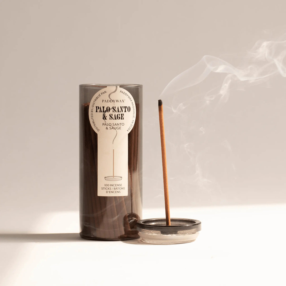 A lit incense stick in a small holder releases smoke beside a brown cylindrical Palo Santo + Sage - Haze Incense container of 100 sticks, making an elegant addition to any incense collection on a light surface with a neutral background.