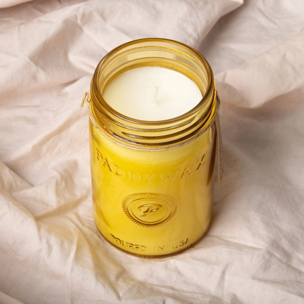 A Fresh Meyer Lemon - Relish 9.5 oz Candle in a yellow Paddywax glass jar with a white wick rests on light, crumpled fabric, ready to brighten your space with its citrus fragrance.