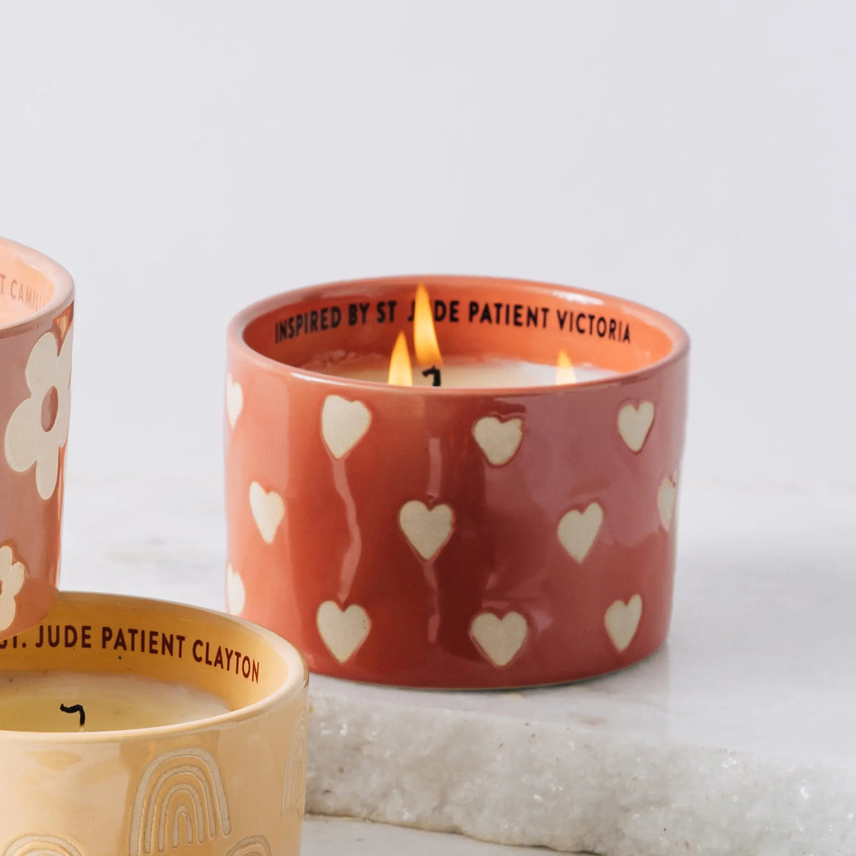 A lit Rosewater Santal - St. Jude Giveback "Courage" 11 oz. Candle with heart designs sits on white marble beside another candle, both supporting the fight against childhood cancer.