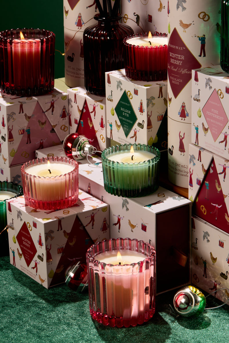 Colorful glass candles, some lit, surrounded by festive boxed packaging with holiday illustrations, arranged on a green surface with Christmas ornaments scattered around.