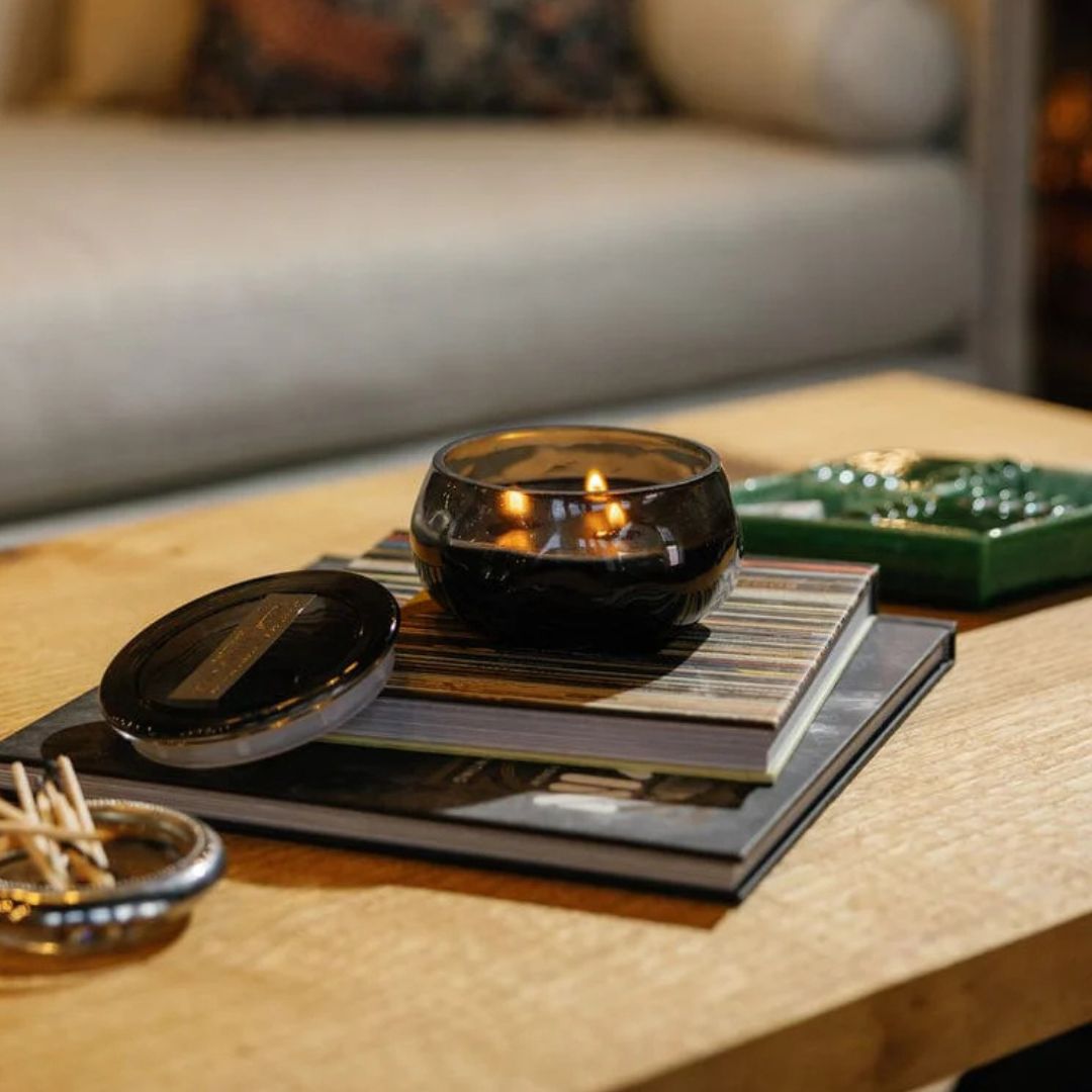 A lit candle in a glass holder sits on stacked books atop a wooden table, with matches beside it and a sofa in the background.