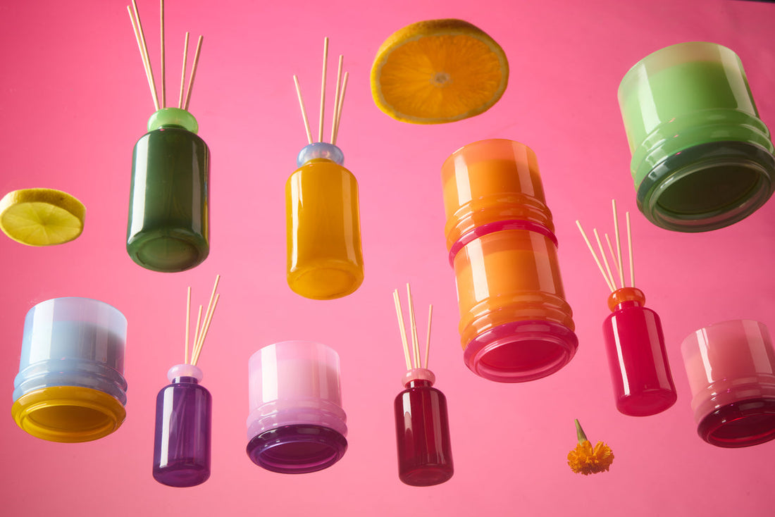 Colorful glass diffuser bottles with wooden sticks and floating slices of citrus fruit and a flower are arranged against a pink background, creating a vibrant, playful composition.