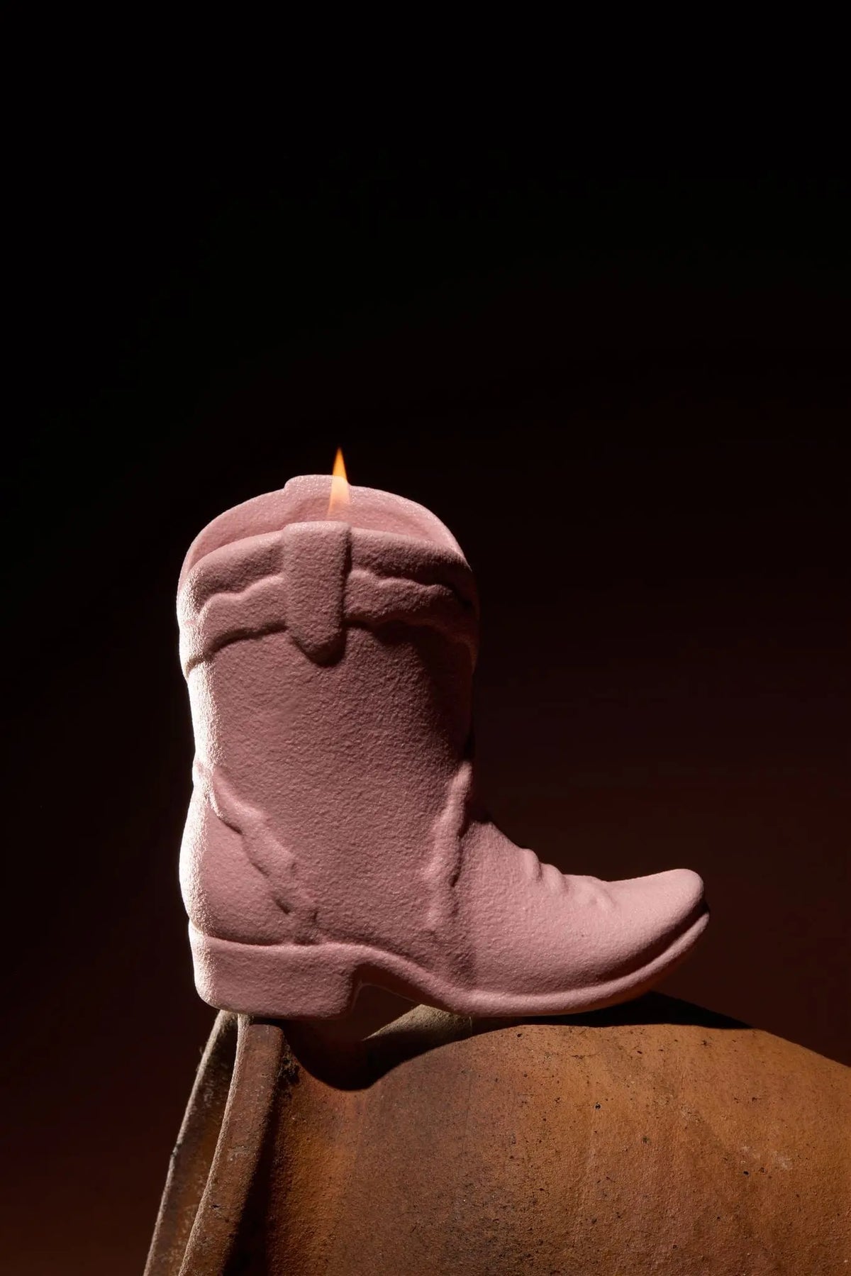 The Yuzu Woods - Nashville Boot 6 oz Candle, shaped like a pink cowboy boot and inspired by Nashville's charm, rests on a curved brown surface with its wick alight and a small flame flickering at the top against a dark background.