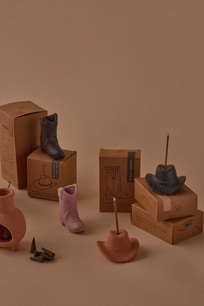 Teak & Tobacco - Incense Holders, including cowboy boots and hats, some with incense sticks, are arranged on and around cardboard boxes against a beige background.