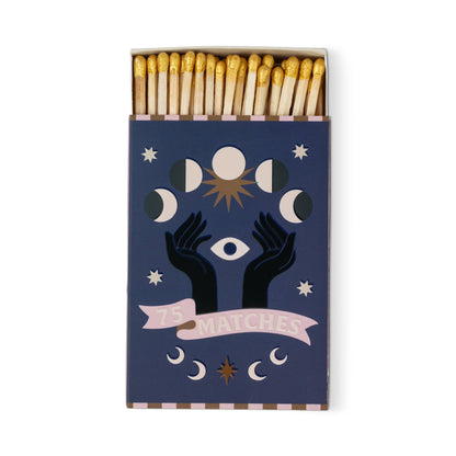 A Dopo "Moon" - 75ct Boxed Matches: Gold-tipped safety matches in a dark blue box with mystical designs—two black hands, an eye, moon phases, stars, and a pink "75 MATCHES" banner. Ideal for lighting your A Dopo candle or bringing Mediterranean vibes.