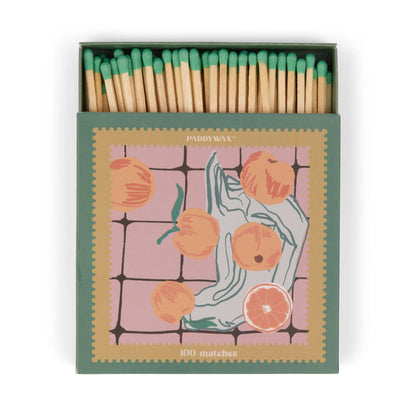 A box of 100 jumbo green-tipped matches, "Oranges - Green Matches," comes in an artful matchbox adorned with peaches and sliced citrus on a pink-tiled background—a stylish candle accessory.