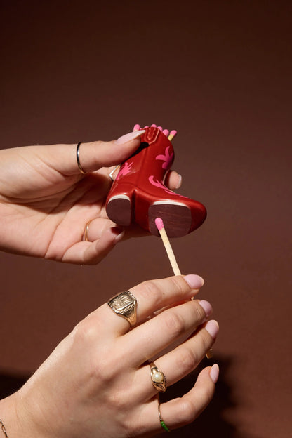 A person holds the Cowboy Boot Match Holder - Red & Pink Flame, a small ceramic piece featuring red and pink details, and uses a cotton swab to clean its sole; the background is brown, and rings on both hands add a charming touch of Western home decor.