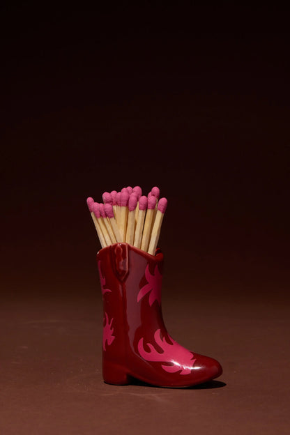 The Cowboy Boot Match Holder - Red & Pink Flame is a small red ceramic boot that holds a bunch of wooden matches with pink tips, set against a plain brown background—perfect for adding a touch of Western home decor or as a unique Nashville gift.