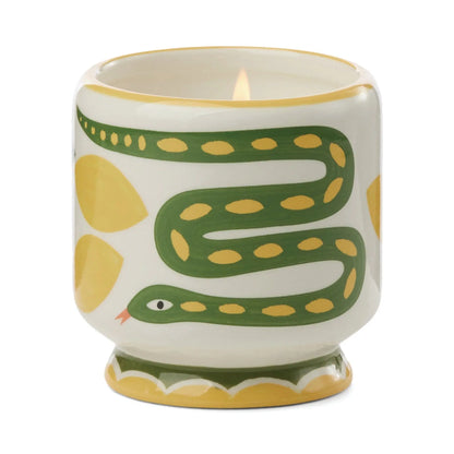 The Wild Lemongrass - A Dopo 8 oz Candle is a lit ceramic candle in a holder with green snake and yellow designs on white, blending artistic charm and Mediterranean fragrance.