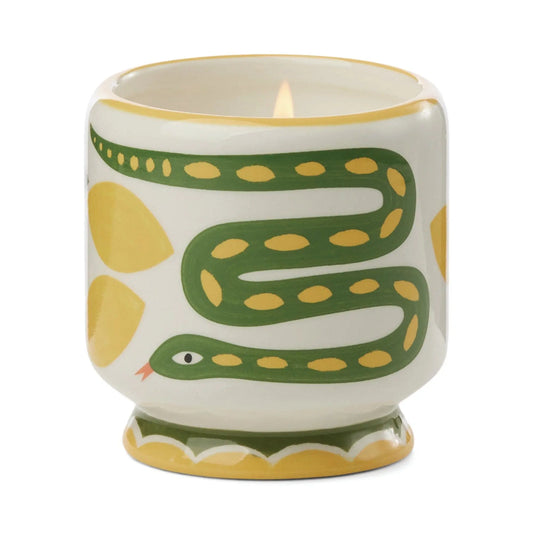 The Wild Lemongrass - A Dopo 8 oz Candle is a lit ceramic candle in a holder with green snake and yellow designs on white, blending artistic charm and Mediterranean fragrance.