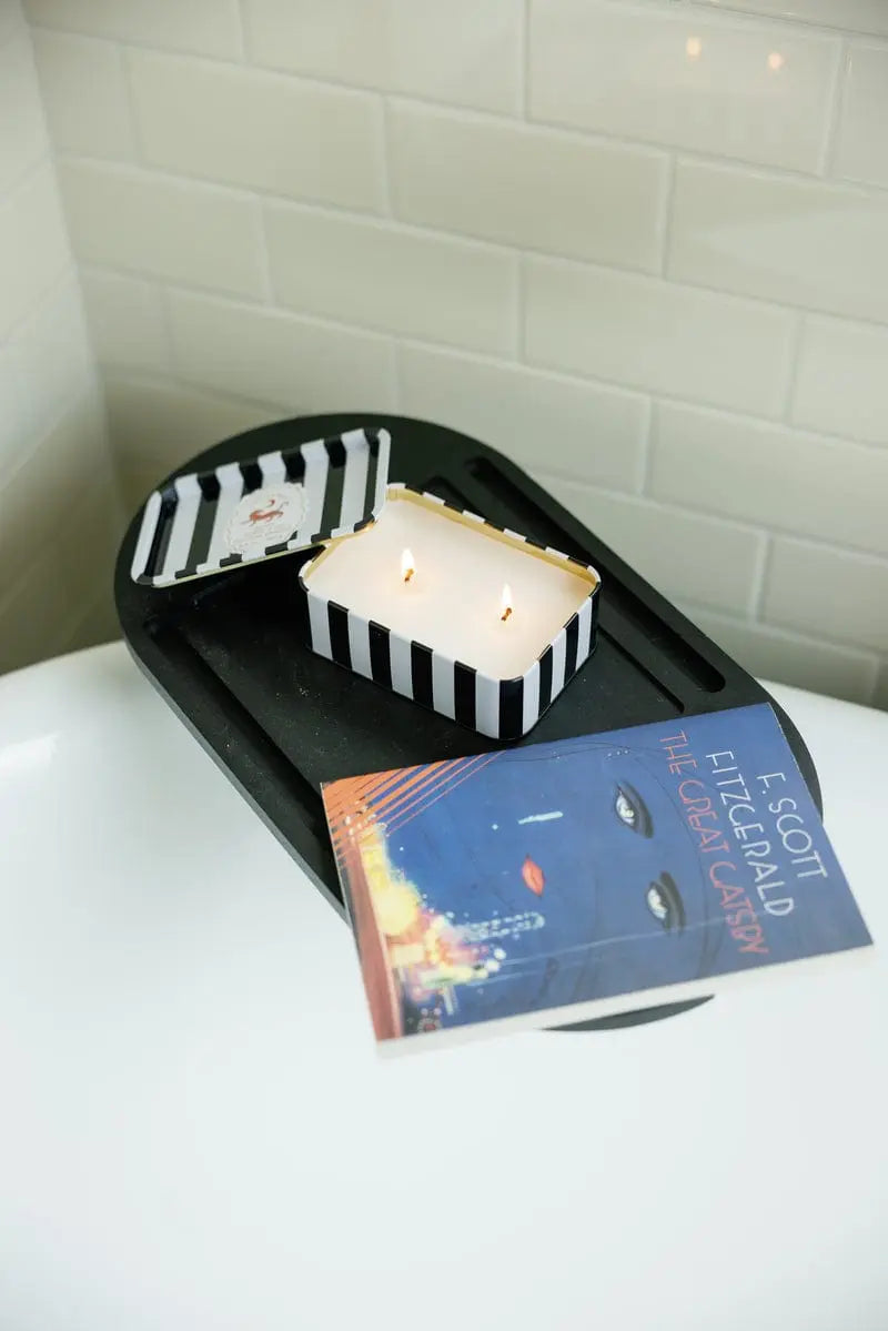 A Black Cedar & Fig - A Dopo 6.5 oz Tin Candle and a copy of The Great Gatsby rest on a black tray above a white bathtub, with white subway tile in the background.