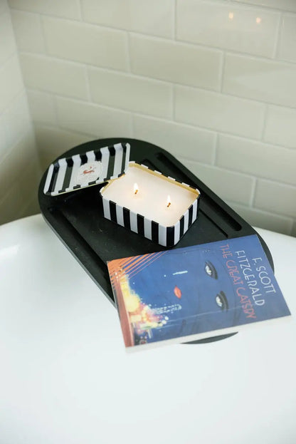 A Black Cedar & Fig - A Dopo 6.5 oz Tin Candle and a copy of The Great Gatsby rest on a black tray above a white bathtub, with white subway tile in the background.