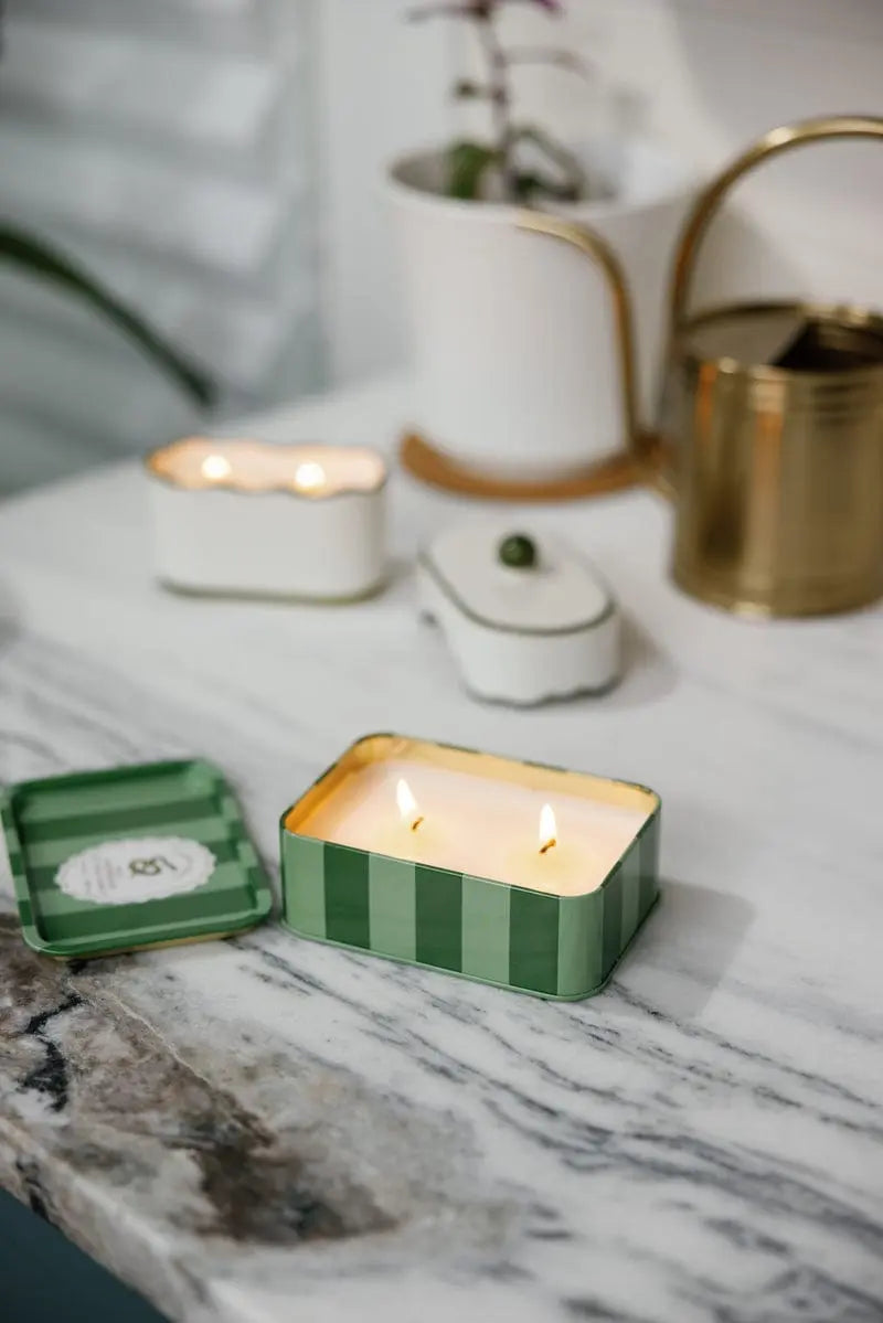 The Wild Lemongrass - A Dopo 6.5 oz Tin Candle, featuring green stripes and a rectangular tin with two lit wicks, rests on a marble surface. In the background, a gold watering can, potted plant, and white containers add decorative touches.