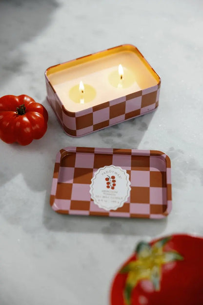 A rectangular Heirloom Tomato - A Dopo 6.5 oz Tin Candle, featuring a pink and red checkered reusable tin with its lid off to reveal the label, sits on a marble surface with two wicks alight; nearby rests a red tomato decoration, evoking the fresh charm of an heirloom tomato.