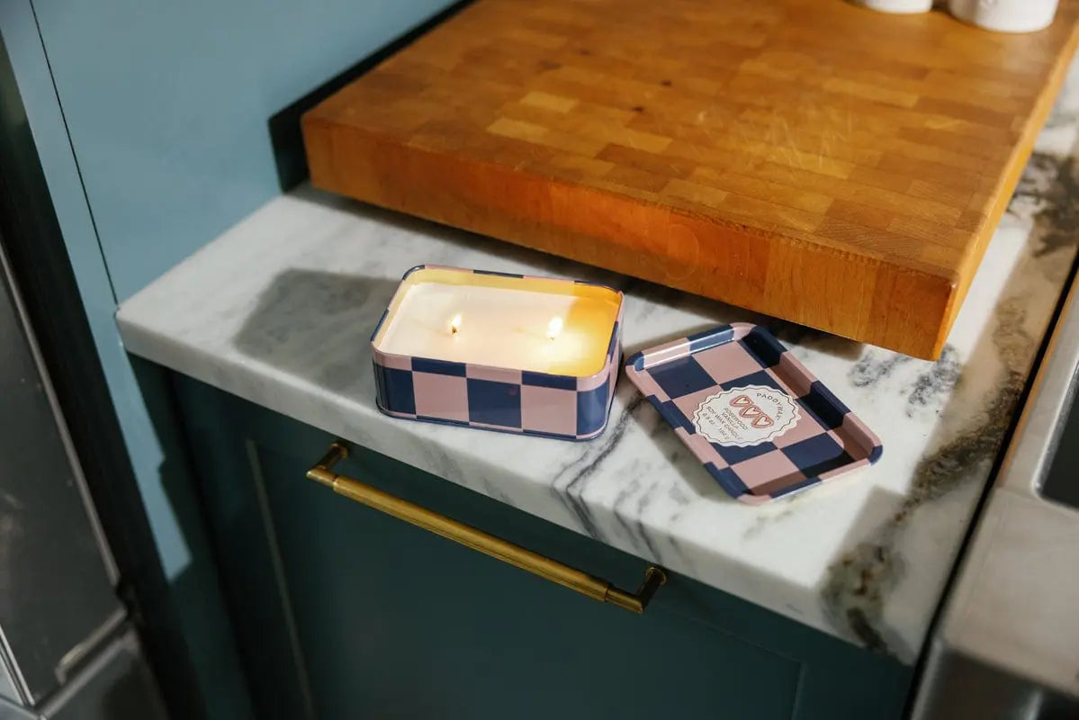 A lit Rosewood Vanilla - A Dopo 6.5 oz Tin Candle, renowned for its bold Mediterranean-inspired fragrance, rests in its open checkered tin on a marble counter beside its lid, beneath a large wooden cutting board.