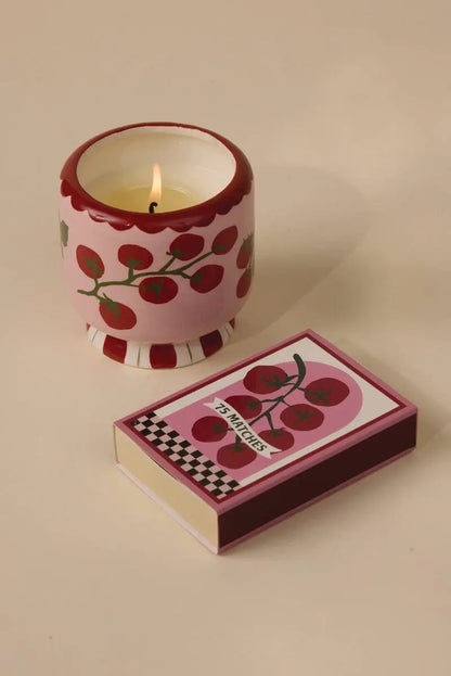 An A Dopo candle in a pink holder with red cherry designs sits next to an A Dopo "Tomato Vine" 75ct boxed matches, both sharing the same cheerful pattern reminiscent of Mediterranean travel, on a beige surface.