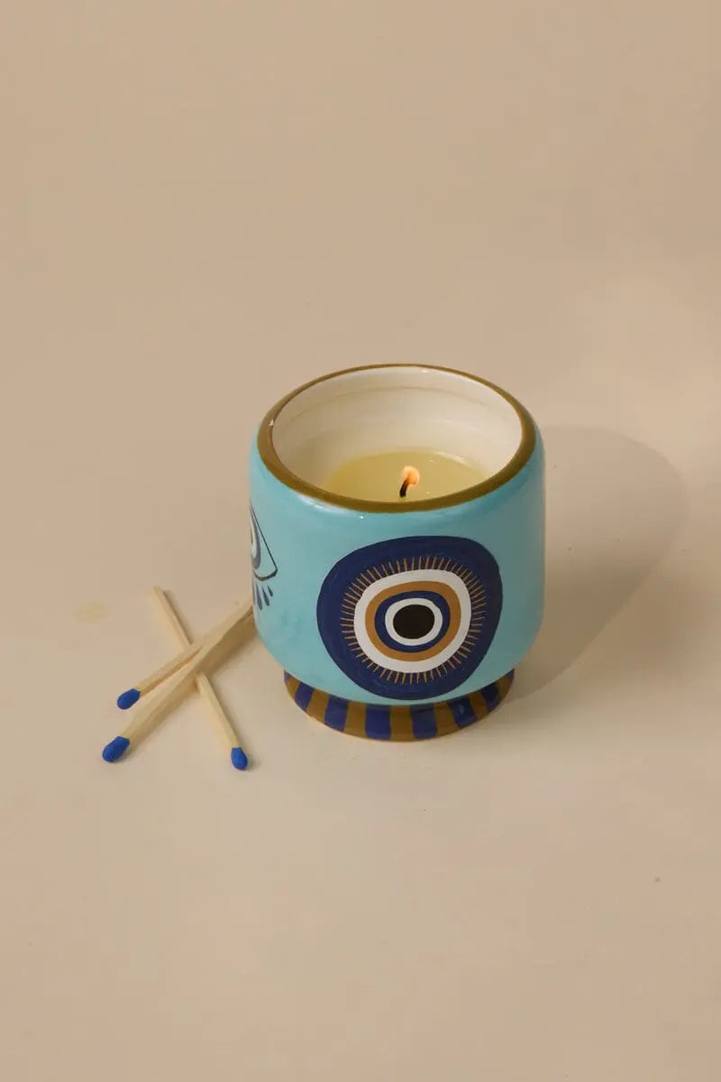 The Incense & Smoke - A Dopo 8 oz Candle in a blue ceramic holder with an eye design sits next to four blue-tipped matches on a light beige surface.