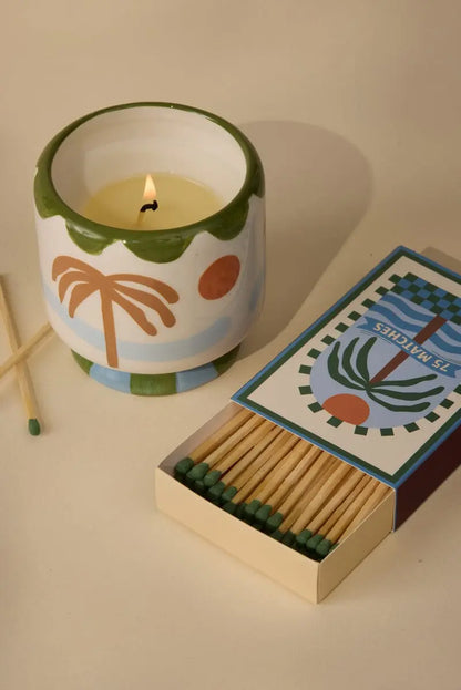 A Dopo Palm Tree 75ct boxed matches sit next to a decorative candle in a ceramic holder with a palm tree motif on a beige surface, evoking memories of Mediterranean travels.